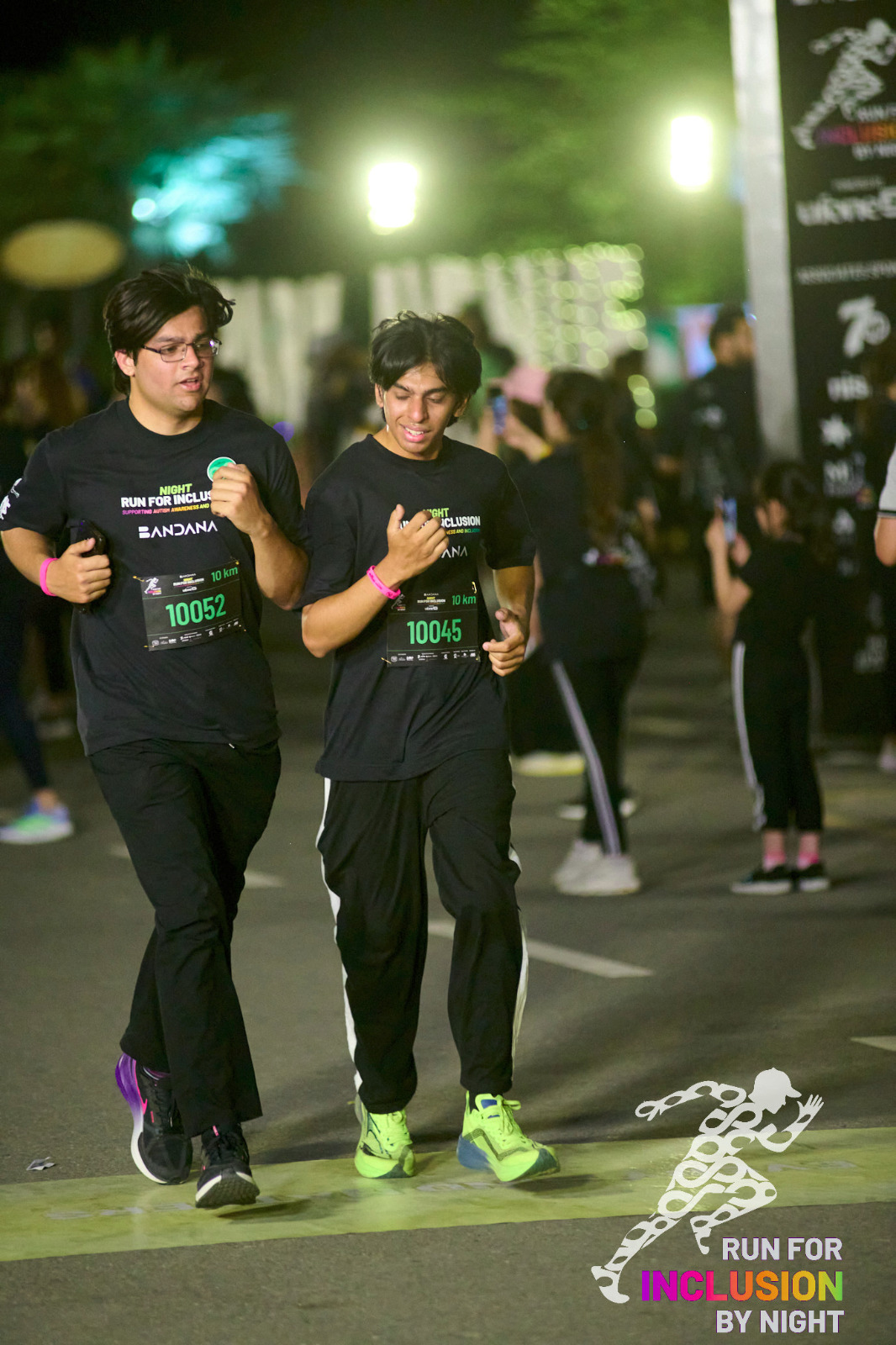 Night race, both of us in black Run for Inclusion shirts, bibs 10052 and 10045, doubled over.